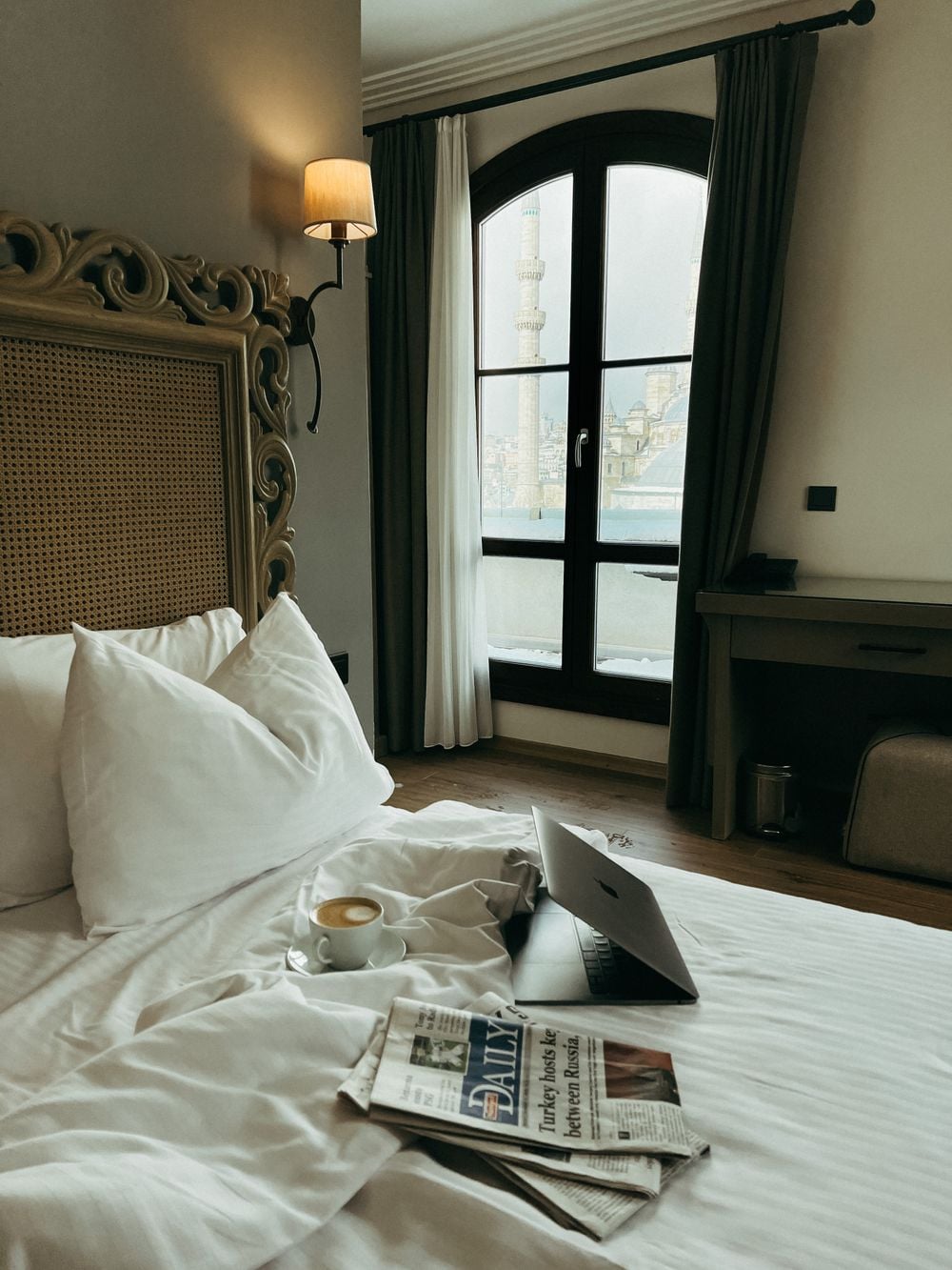 A cozy hotel room with an unmade bed, a laptop, a newspaper, and a cup of coffee. A large window lets in natural light and offers a view of a mosque with minarets outside.