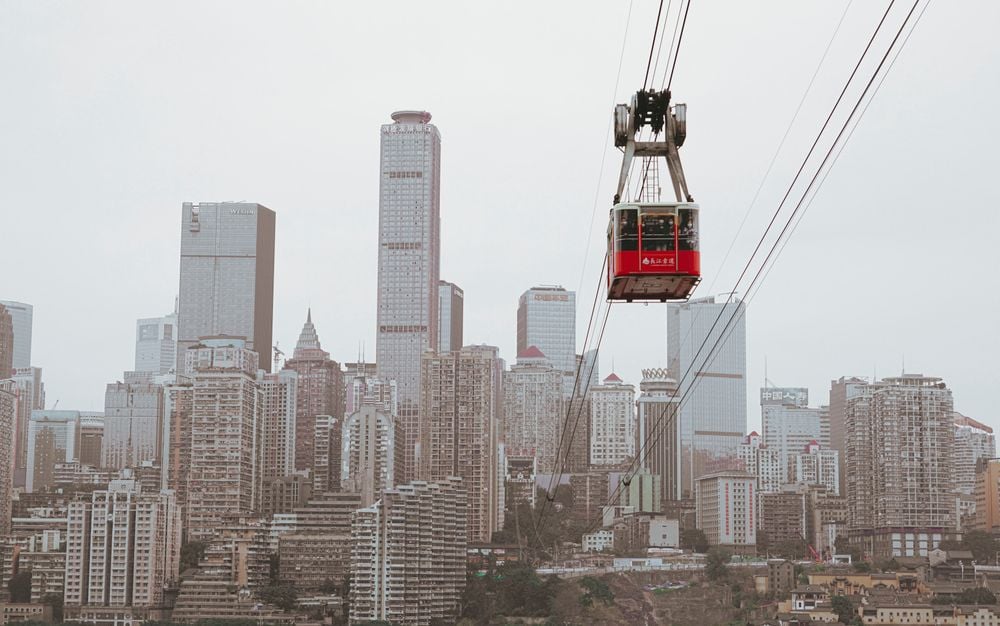 A red cable car travels above a dense cityscape with numerous tall buildings and skyscrapers on a cloudy day.