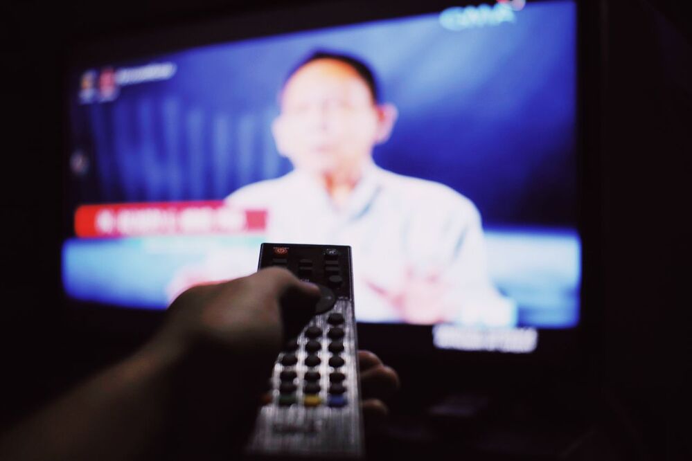 A hand holding a remote control is pointed at a TV screen displaying a blurred image of a person speaking, with text and graphics partially visible.