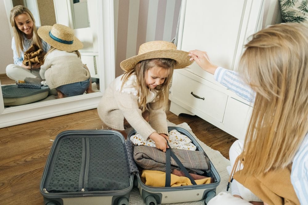 A young girl wearing a straw hat packs clothes into an open suitcase while a woman beside her adjusts the hat. Their reflection is visible in a large mirror behind them.