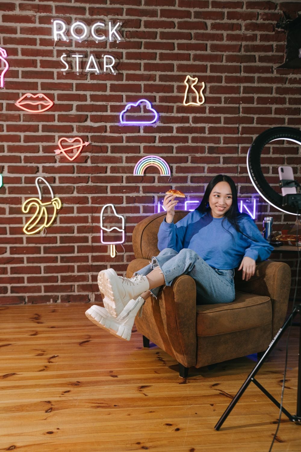 A woman in a blue sweater sits on a brown chair holding pizza. Neon signs and a brick wall are in the background. A ring light stands nearby.