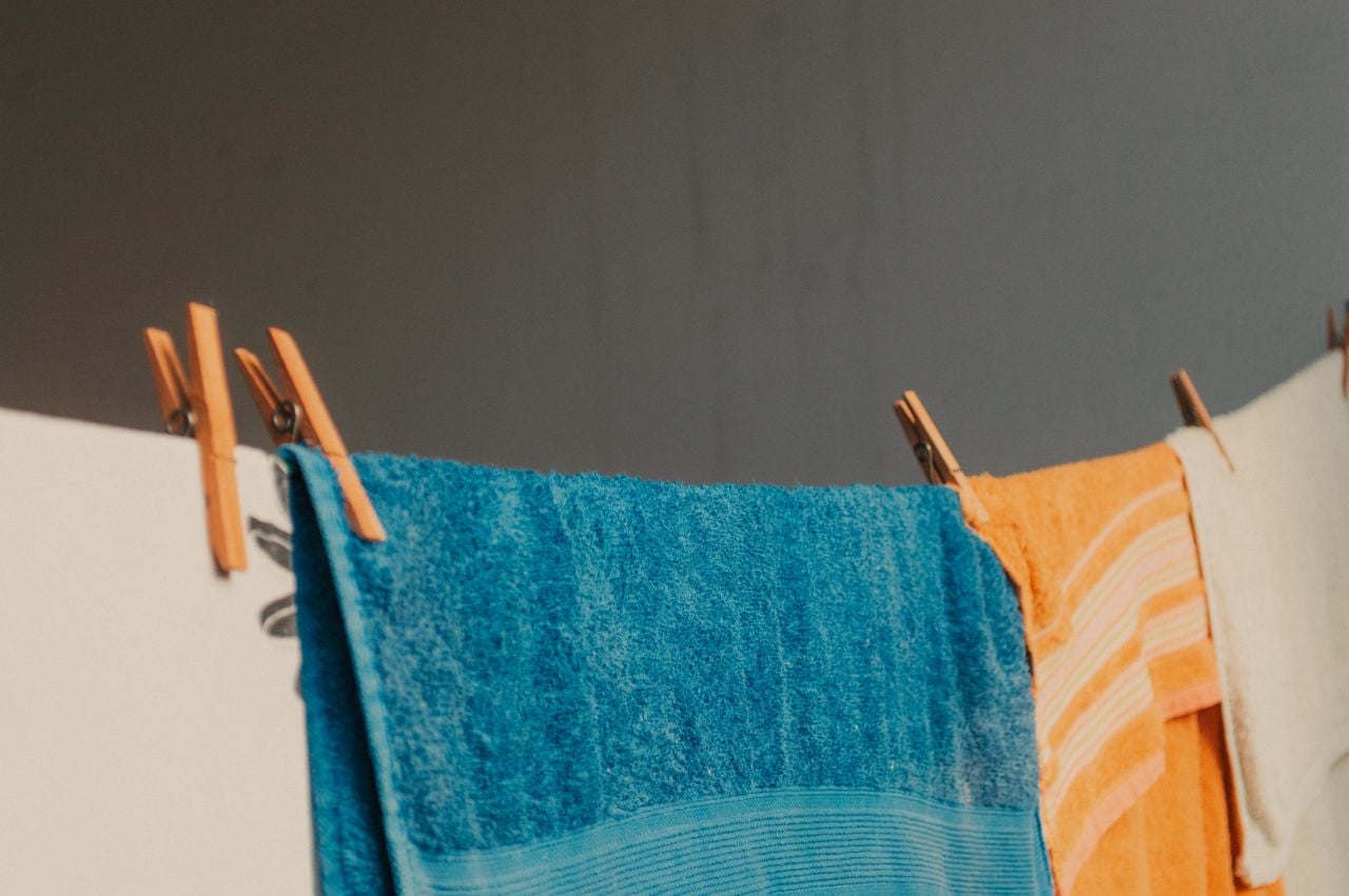 Three towels, one blue, one orange with stripes, and one white, hang on a clothesline with wooden clothespins against a plain gray background.