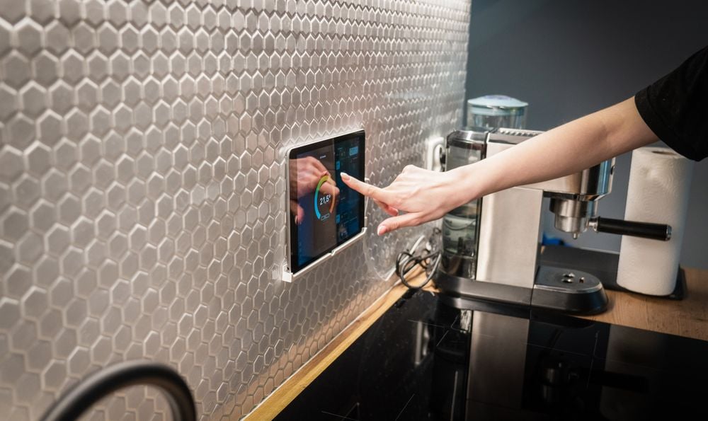 A person touches a wall-mounted touchscreen in a modern kitchen with metallic hexagon tiles, next to a coffee machine and paper towel roll.