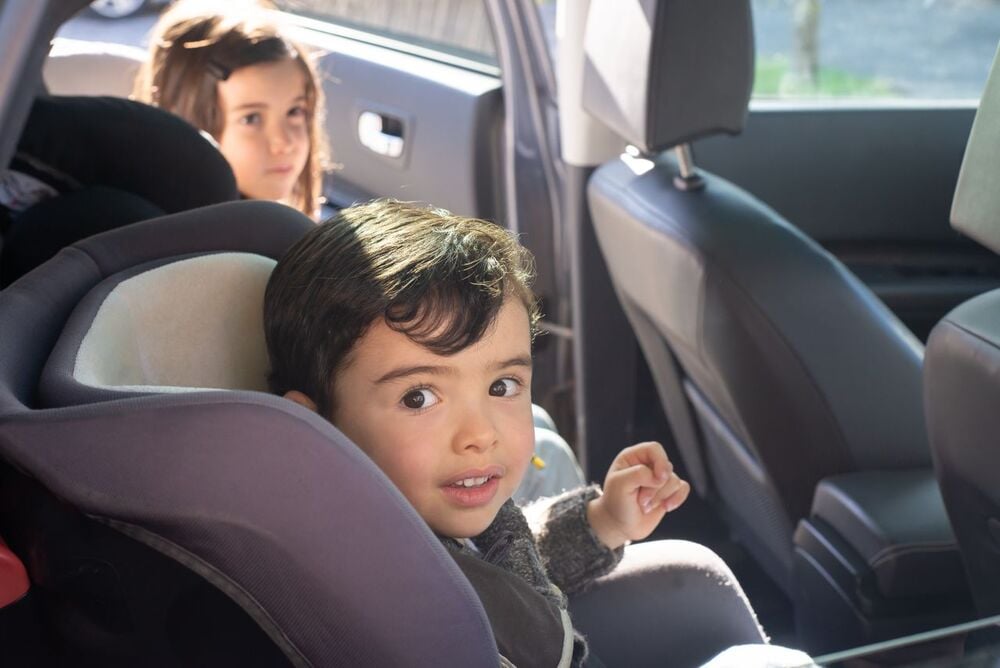 A young child in a car seat looks at the camera, while an older child sits in the back seat, gazing out the window. Sunlight softly illuminates the car interior.