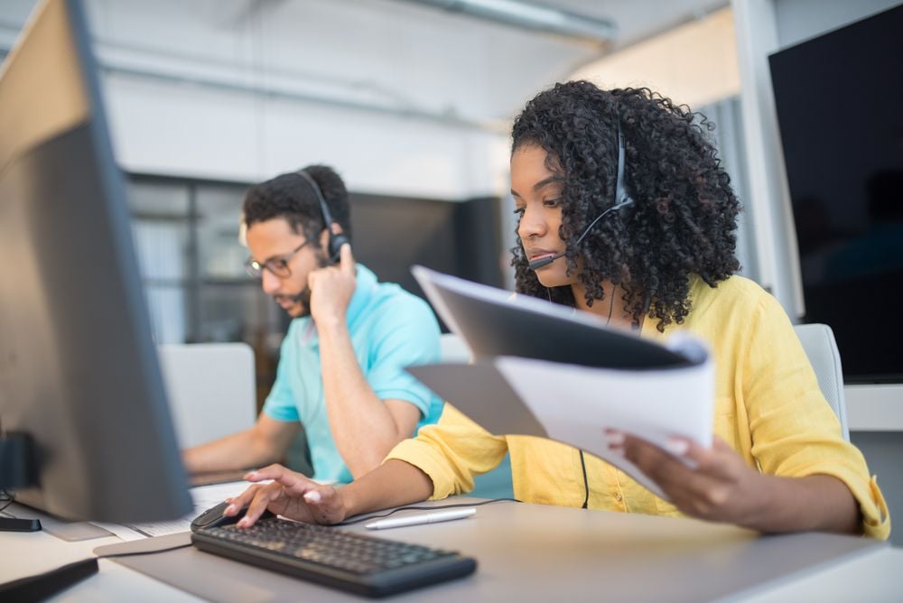 Two people wearing headsets work at computers in an office. The woman in front is holding papers and typing, while the man in the background looks at his screen. Both appear focused on their tasks.