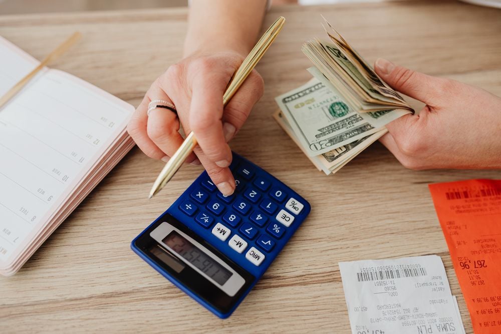 A person uses a blue calculator and holds a stack of dollar bills, with receipts, a pen, and an open planner on a wooden table.