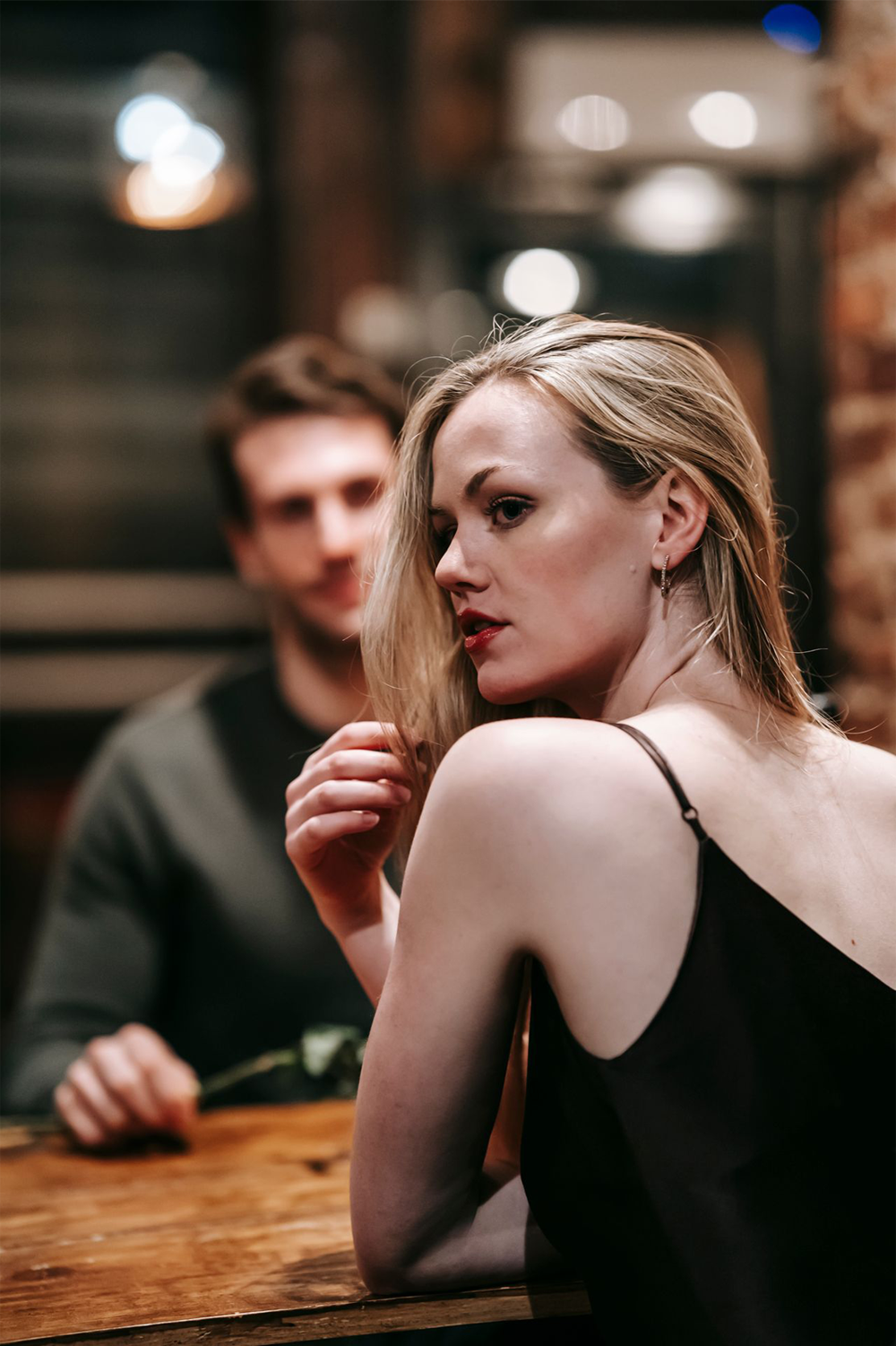 A woman in a black dress sits at a wooden table, looking over her shoulder at the camera, with a man sitting in the blurred background.