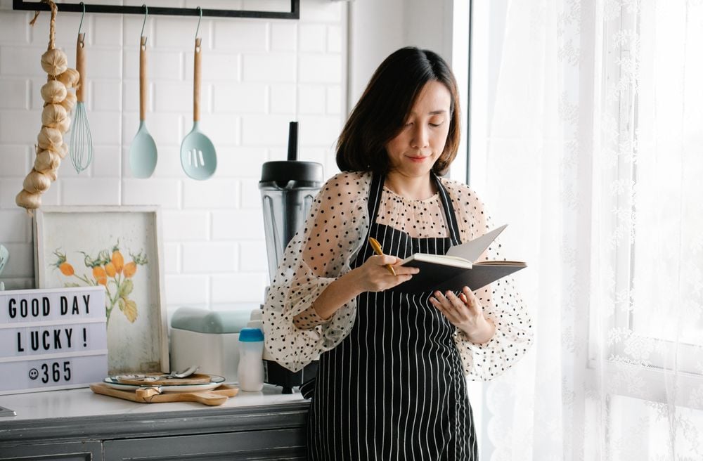A woman in a striped apron writes in a notebook while standing in a bright kitchen. Cooking utensils hang on the wall, and a lightbox reads “GOOD DAY LUCKY! @365” on the counter.
