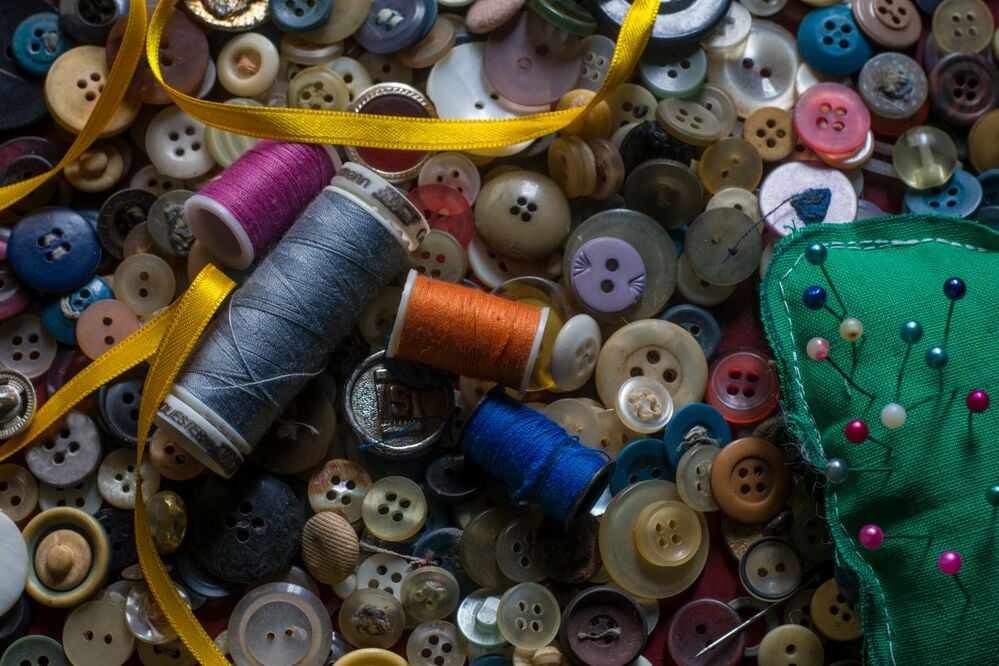 Colorful buttons, spools of thread, yellow ribbon, and a green pincushion with pins are scattered together, creating a busy and vibrant sewing scene.