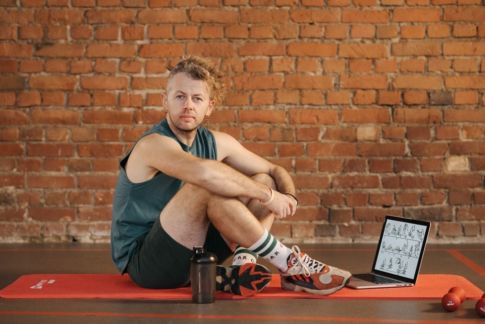 A man sits on a red exercise mat in a gym, next to a laptop displaying workout diagrams, a water bottle, and a small dumbbell, with a brick wall in the background.