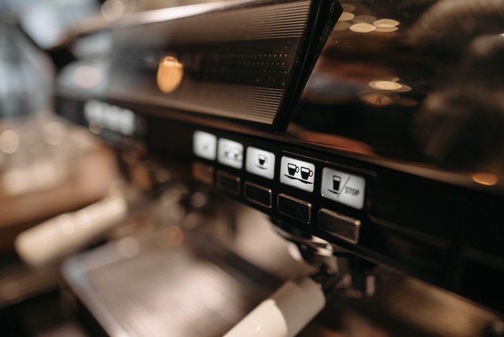 Close-up of a coffee machine control panel with illuminated buttons, including icons for different coffee options and a stop button. The machine is metallic with a modern design, and the background is softly blurred.