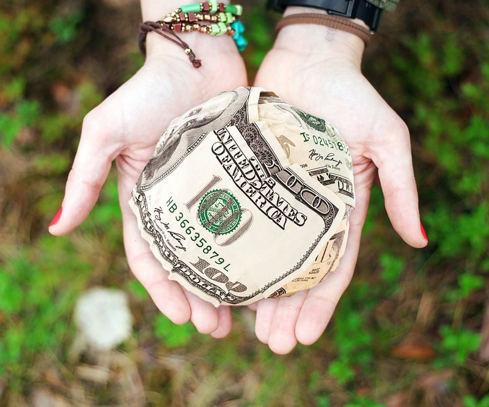 Two hands gently hold a round object made of crumpled U.S. dollar bills, with greenery visible in the blurred background.