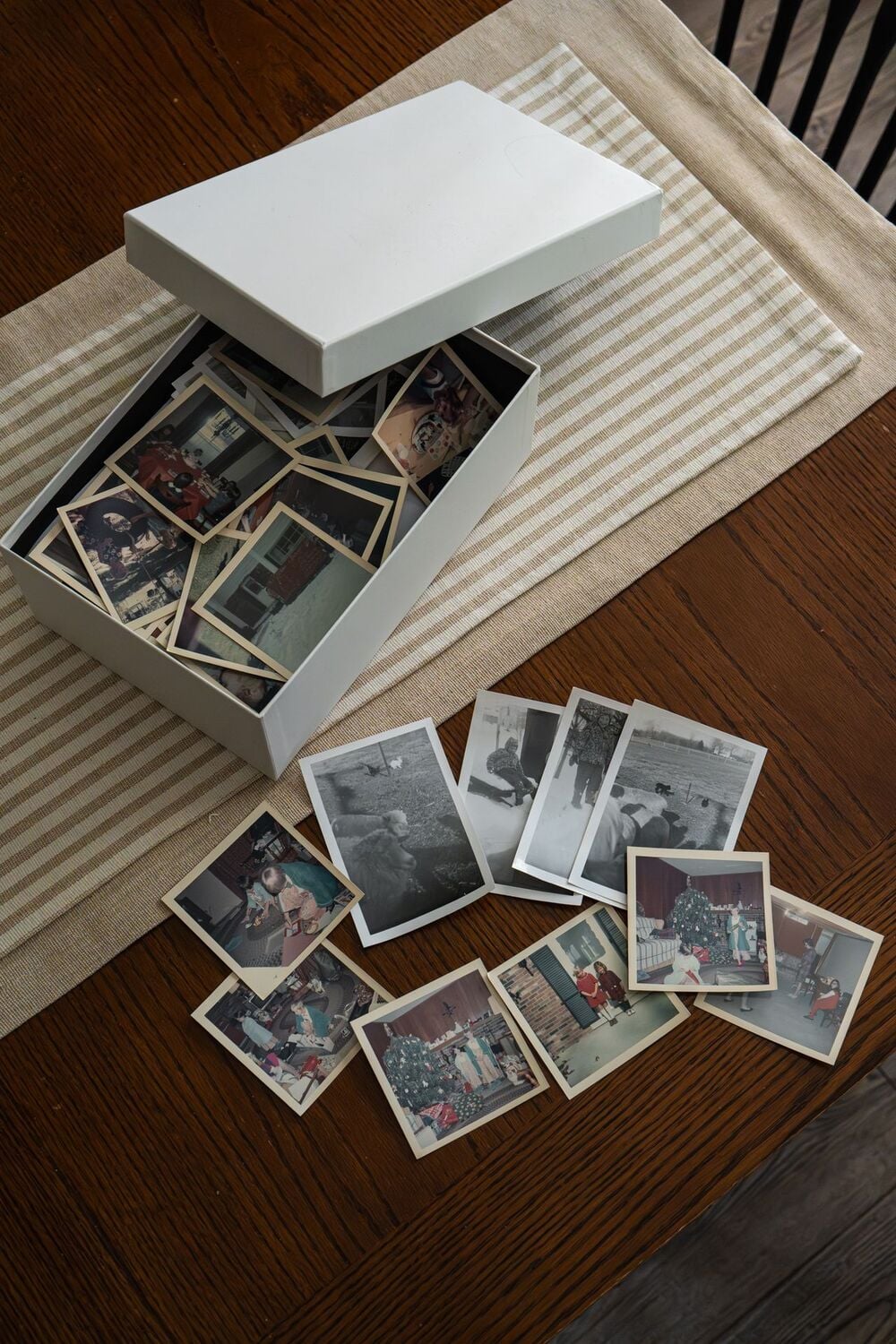 A white box filled with old photographs sits on a wooden table. Several vintage photos, some black and white and some color, are spread out on the table next to the box.