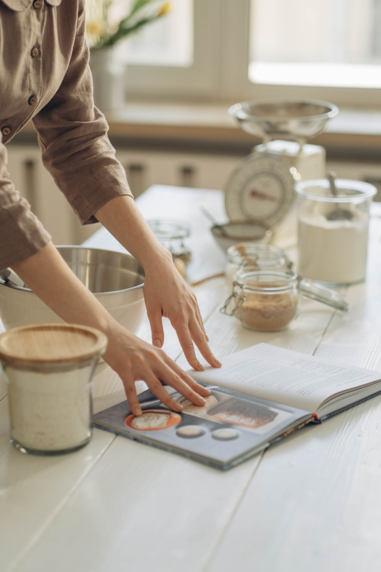 A person in a brown shirt prepares to bake, referencing an open cookbook on a white kitchen table with jars of ingredients, a mixing bowl, and a kitchen scale nearby.