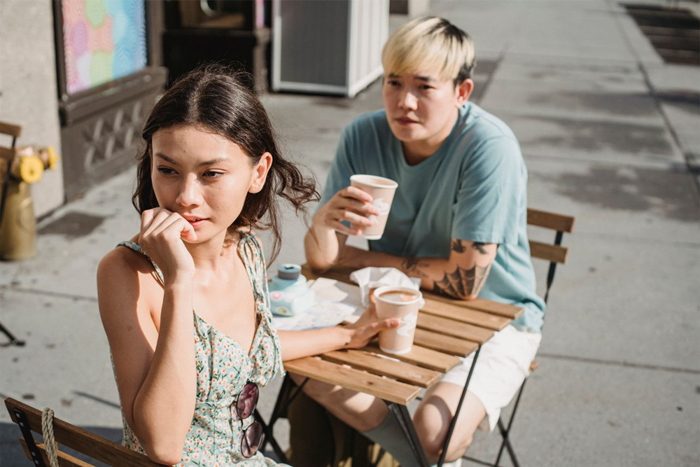 Two people sit at a small outdoor cafe table with coffee cups. The woman in front looks away pensively, while the man behind her gazes in her direction, holding his cup. It’s a sunny day.