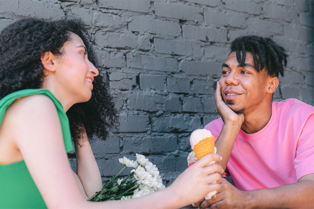 Two people sit together by a gray brick wall. One holds a bouquet of white flowers and an ice cream cone. They smile at each other, appearing to enjoy a pleasant conversation.