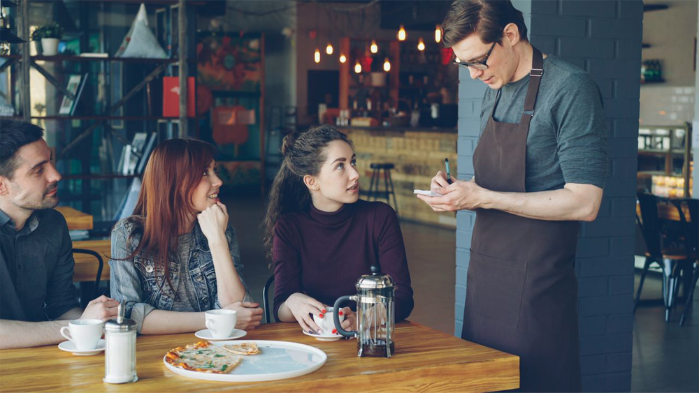A waiter in an apron takes an order from three people seated at a café table with coffee cups, a French press, and pizza. The group looks at the waiter attentively in a cozy, warmly lit setting.