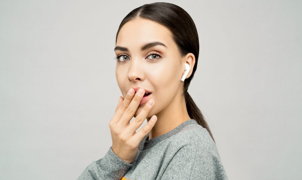 Woman with dark hair and earbuds wears a grey top, covering her mouth with her hand, looking surprised against a plain light background.