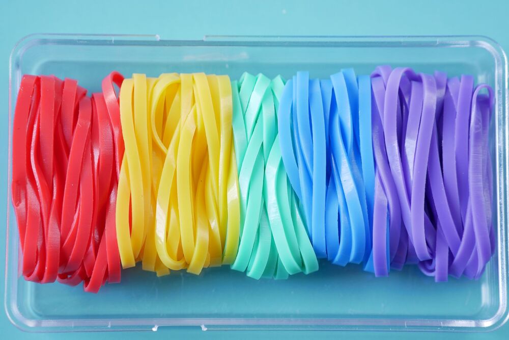 A clear plastic box contains rubber bands arranged by color: red, yellow, green, blue, and purple, displayed in neat rows on a light blue background.