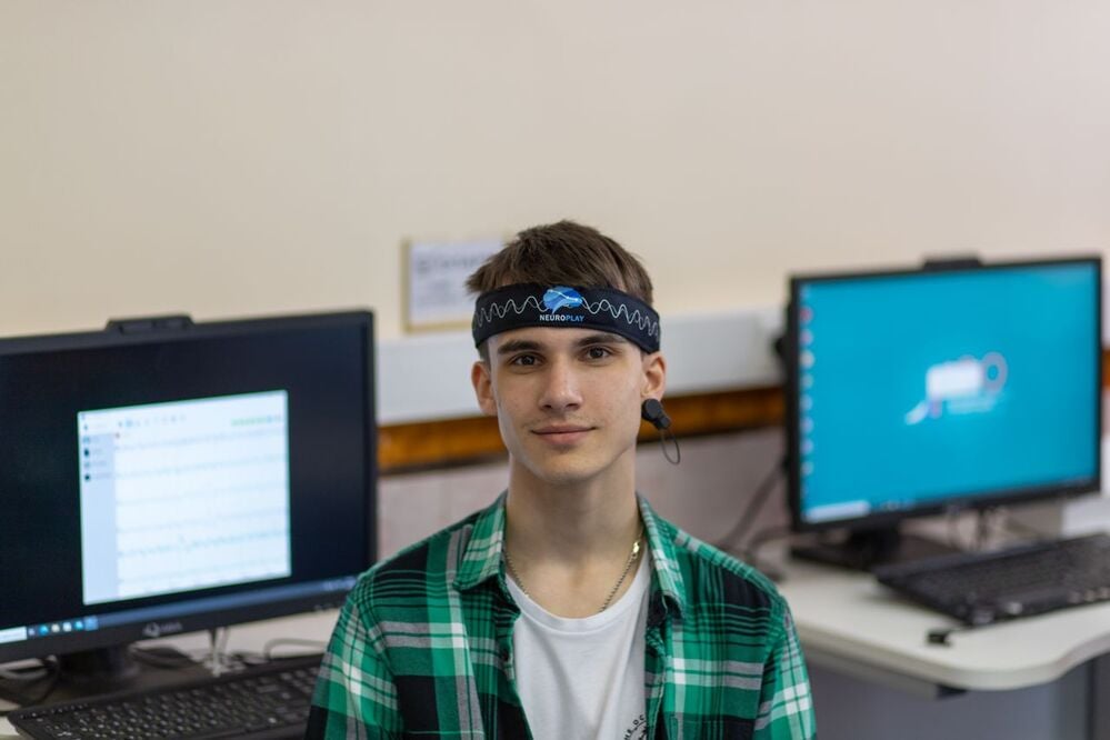 A young person wearing a headband with sensors sits in front of two computer monitors in a classroom or lab setting.