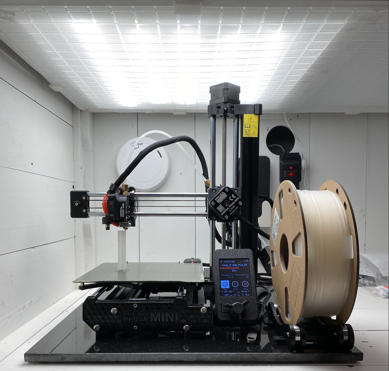 A 3D printer with a spool of filament is set up in a well-lit enclosure, ready for printing. The control screen shows settings, and the walls are white with tools and equipment visible in the background.