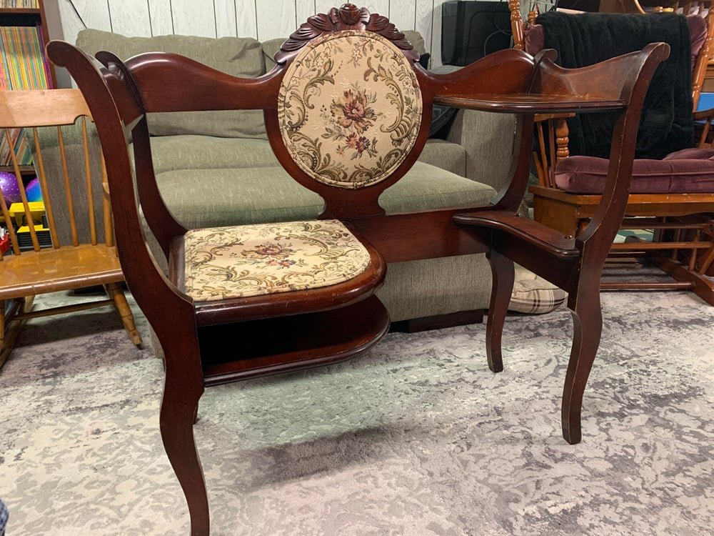 A vintage wooden gossip bench with a floral upholstered seat and backrest, small tabletop surfaces on each side, and curved legs, placed indoors on a patterned rug.