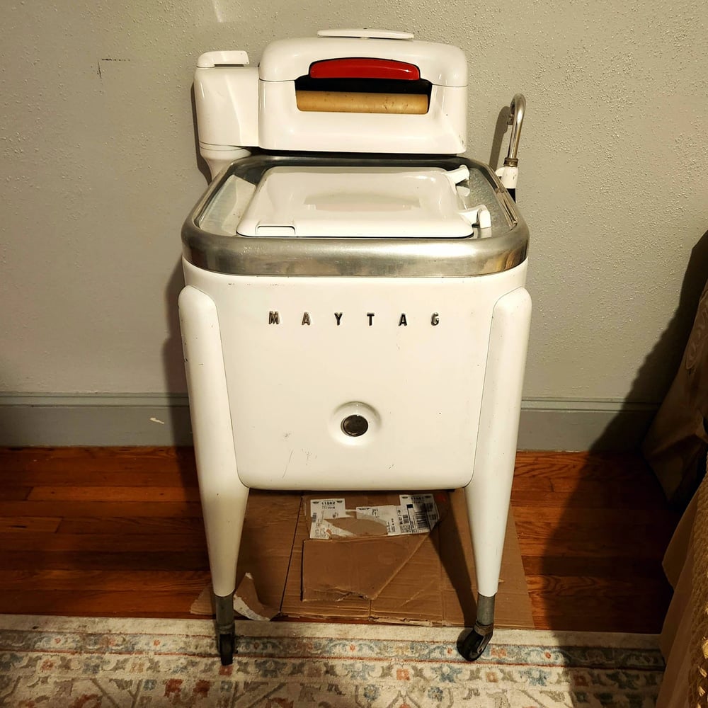 A vintage white Maytag wringer washing machine on wheels stands on a hardwood floor, against a light-colored wall, with a cardboard box underneath.