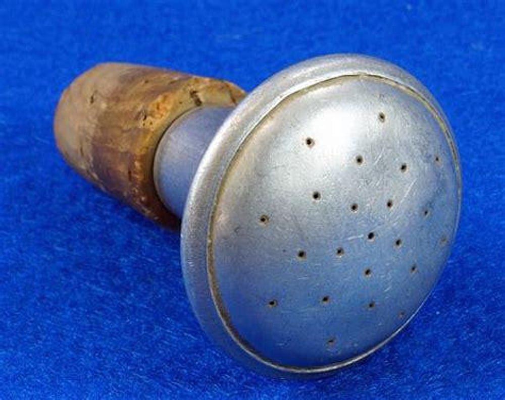 A vintage metal and cork bottle stopper with small holes in the round top, likely used as a shaker or sprinkler, on a blue fabric background.