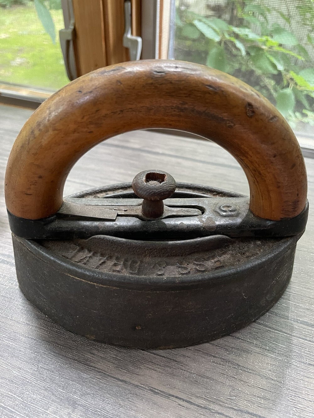 A vintage metal and wood iron sits on a gray surface near a window, with greenery visible outside. The iron has a round wooden handle and a screw on top.