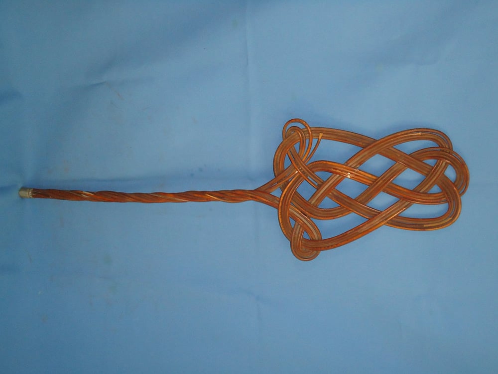 A copper wire is twisted into a looped, intricate knot design at one end, with a straight handle extending from the knot, all on a light blue background.