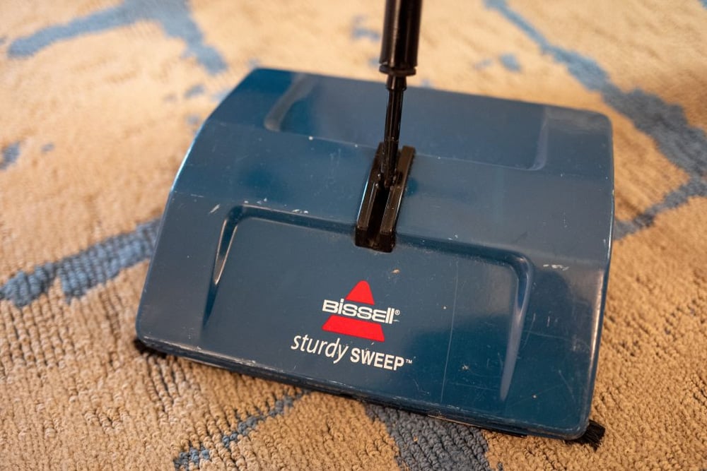 A blue Bissell Sturdy Sweep manual carpet sweeper is shown on a beige carpet with blue patterns.