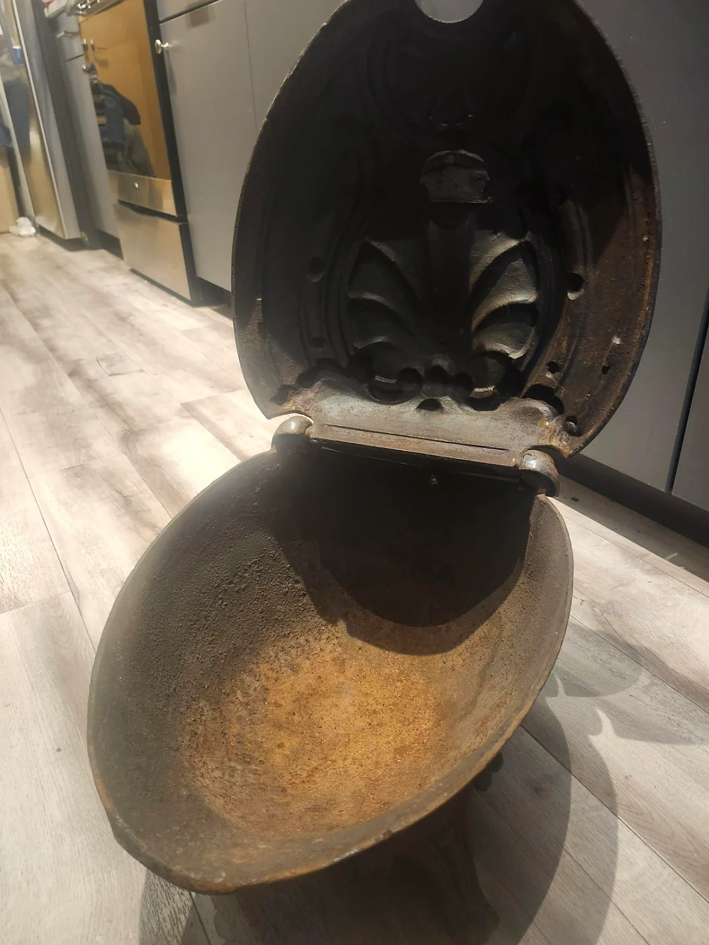 A large, old, oval-shaped cast iron container with a hinged lid, sitting open on a kitchen floor. The interior is rusty and the lid features an ornate, decorative pattern.