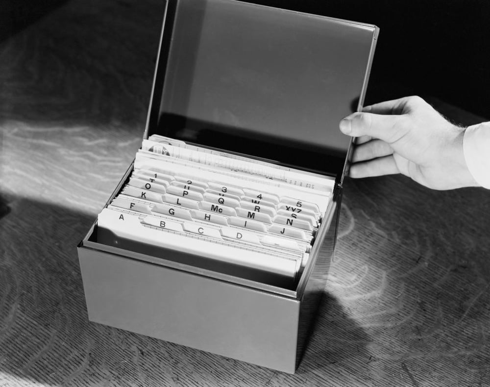 A hand opening a metal box filled with index cards organized alphabetically with dividers, sitting on a wooden surface.