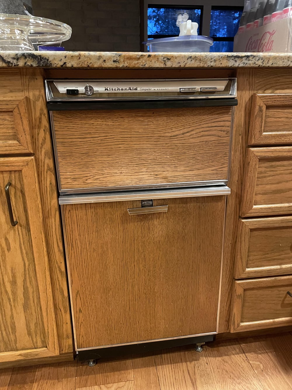 A vintage KitchenAid dishwasher with a wood panel front is built into a wooden kitchen cabinet. A granite countertop is visible above, along with various items and a Coca-Cola box in the background.