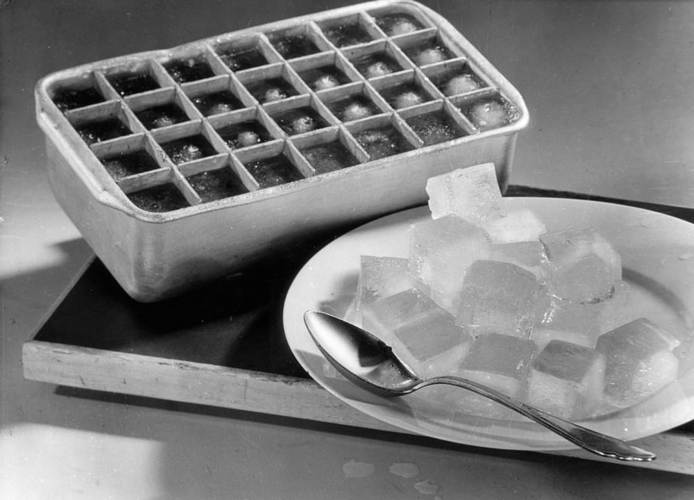 A metal ice cube tray sits next to a white plate with several ice cubes and a metal spoon on it. The scene is set on a table with a simple background.