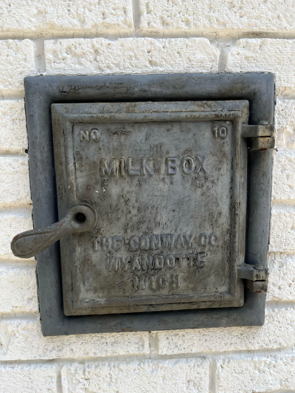 A weathered metal milk box with a handle is set into a white brick wall. The box is labeled “MILK BOX” and has manufacturer and location details embossed on the front.