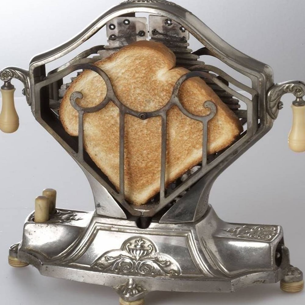 A vintage toaster with ornate metalwork and two slices of toasted bread held upright between metal grates. The toaster features decorative handles and an intricate base design.