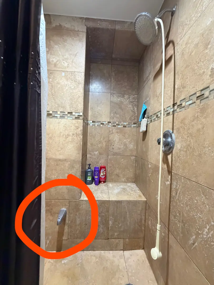 A shower with tan tiles, a built-in bench, several bottles, and a showerhead. A bath spout is oddly placed low on the bench, highlighted by an orange circle.