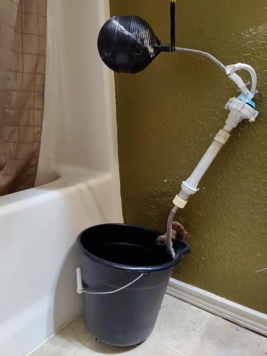 A black bucket on the floor catches water from a homemade device attached to the wall, using pipes and a float valve, next to a bathtub with a beige shower curtain.