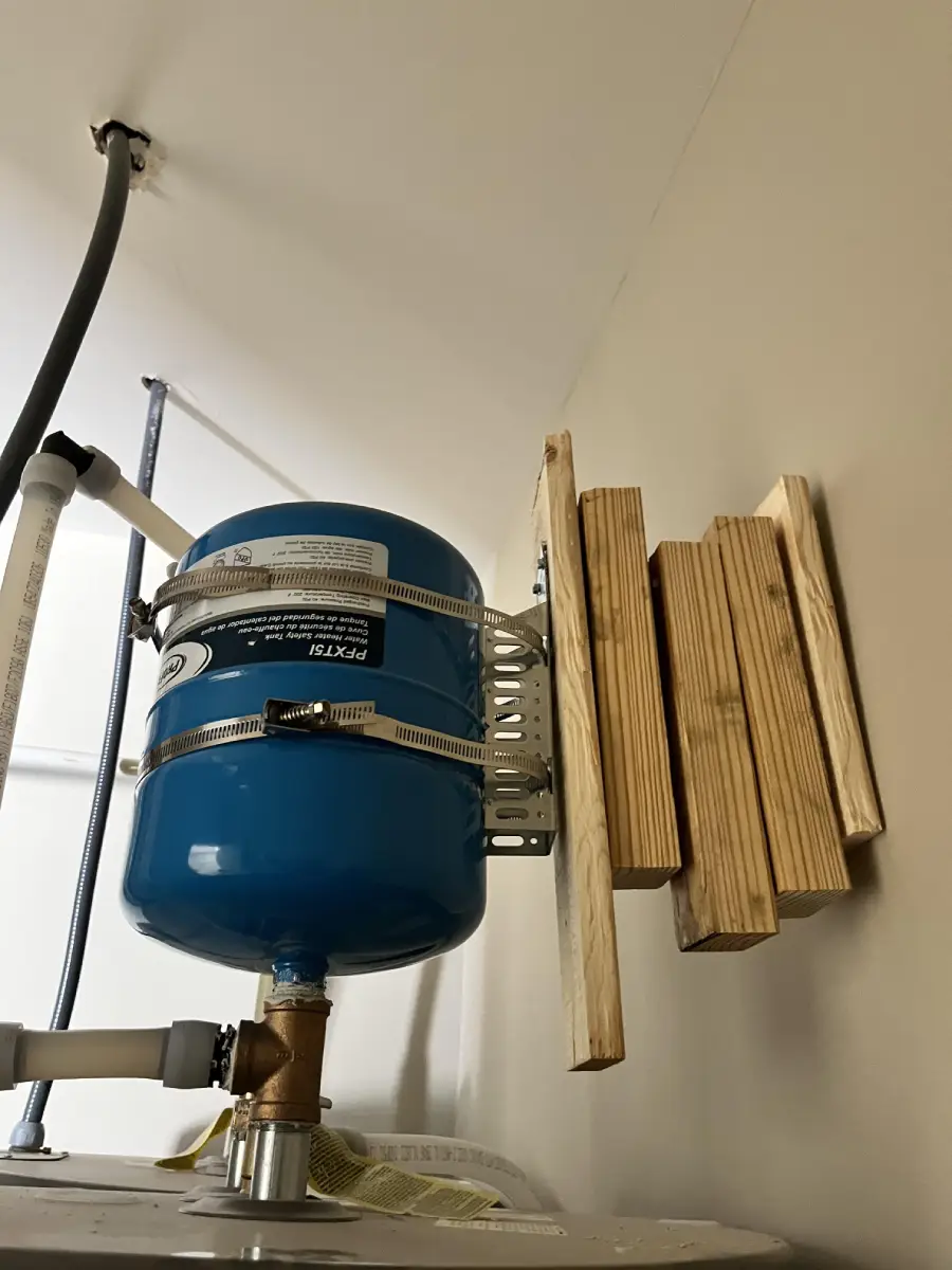 A blue pressure tank is mounted on pipes with metal brackets. Several pieces of cut wood are attached to the wall next to it, creating a makeshift barrier or support. The setup appears to be in a utility room.