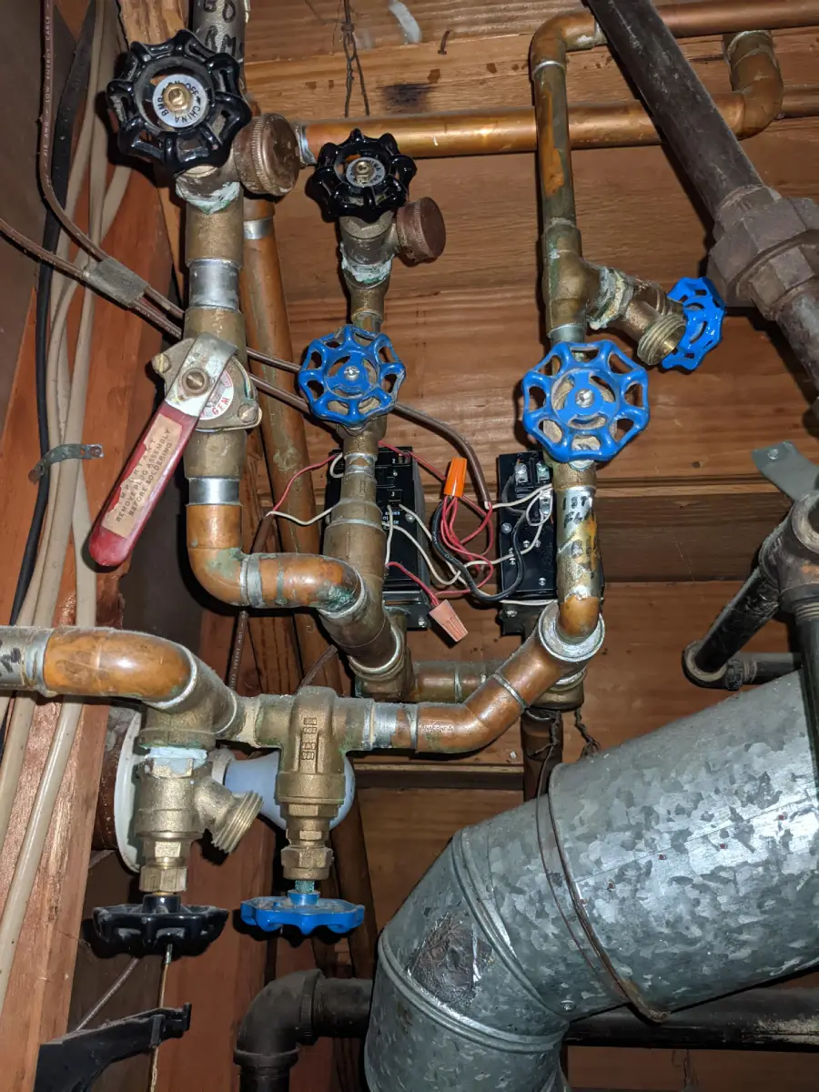 A complex arrangement of copper pipes and valves, some with blue and black handles, is mounted against a wooden wall, with electrical wires and a large metal air duct visible nearby.