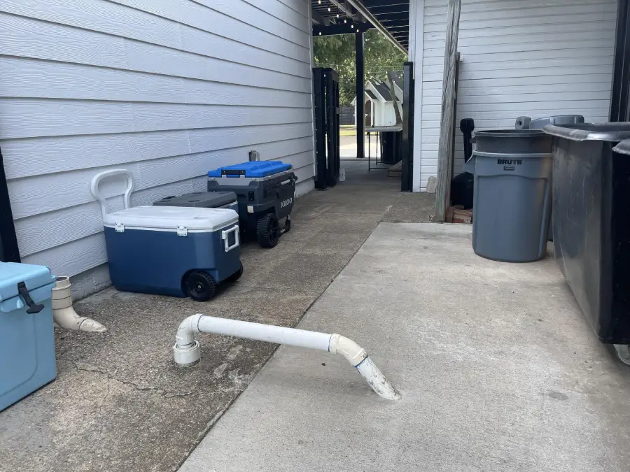 A bent white PVC pipe sticks out of a concrete patio near coolers, trash bins, and a white-paneled building. A narrow walkway leads to an open area with trees in the background.