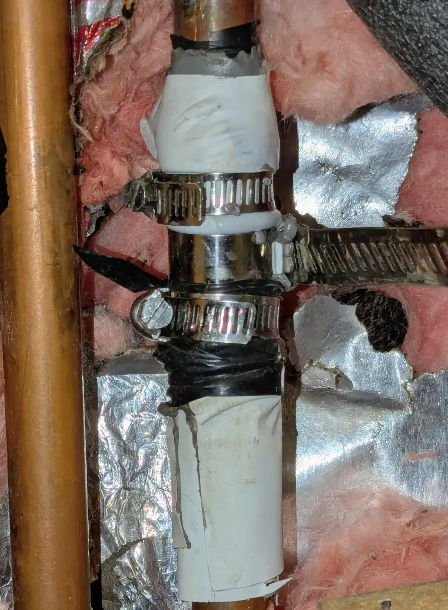Close-up of a pipe repair using white tape, black tape, and several metal hose clamps, surrounded by insulation and reflective material.
