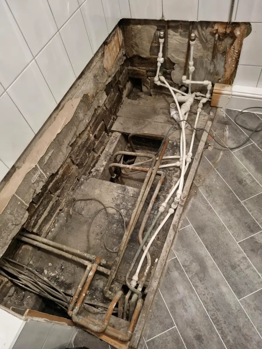 Exposed plumbing and pipes in a bathroom wall and floor, with tiles and part of the wall removed, showing various metal and plastic pipes and fittings.