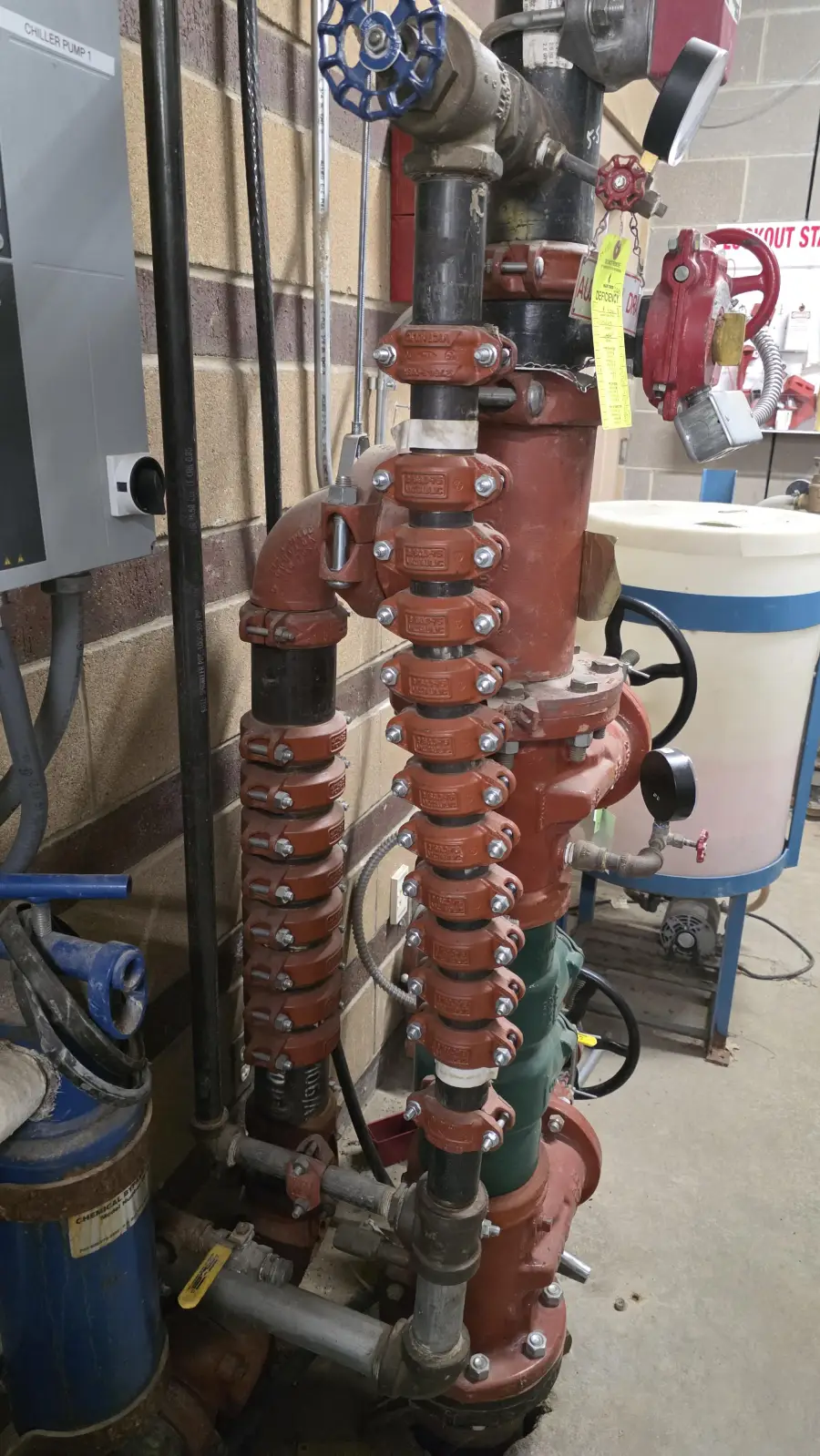 A complex plumbing system with multiple red and black valves, pipes, and fittings, installed against a brick wall inside an industrial or utility room setting.