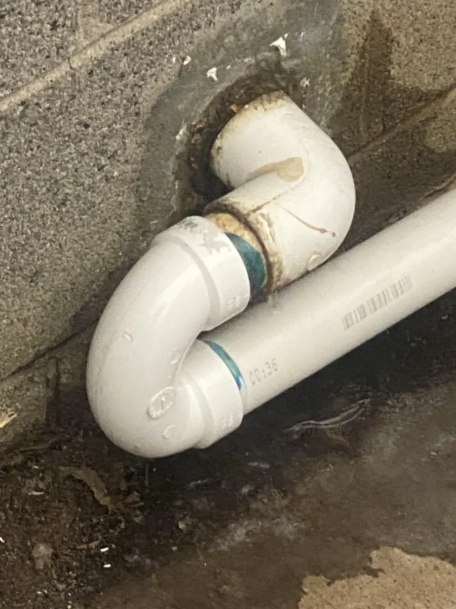 A white PVC pipe is installed very close to a concrete wall, with water stains and droplets visible on the pipe and wet spots on the ground below.