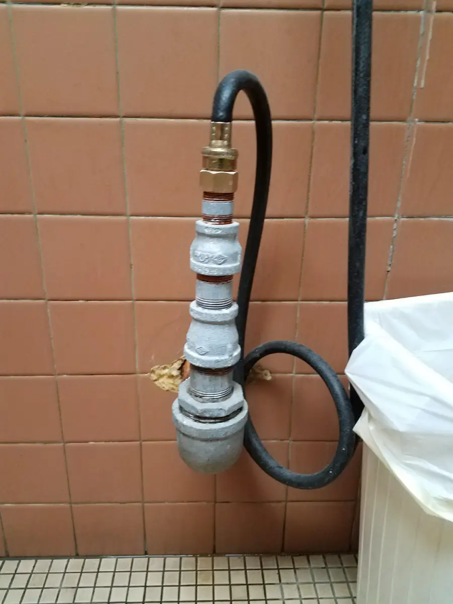 A black hose connected to a series of metal fittings is attached to a tiled wall above a tile floor. Part of a white bin with a liner is visible on the right. Some tiles behind the fittings are cracked.