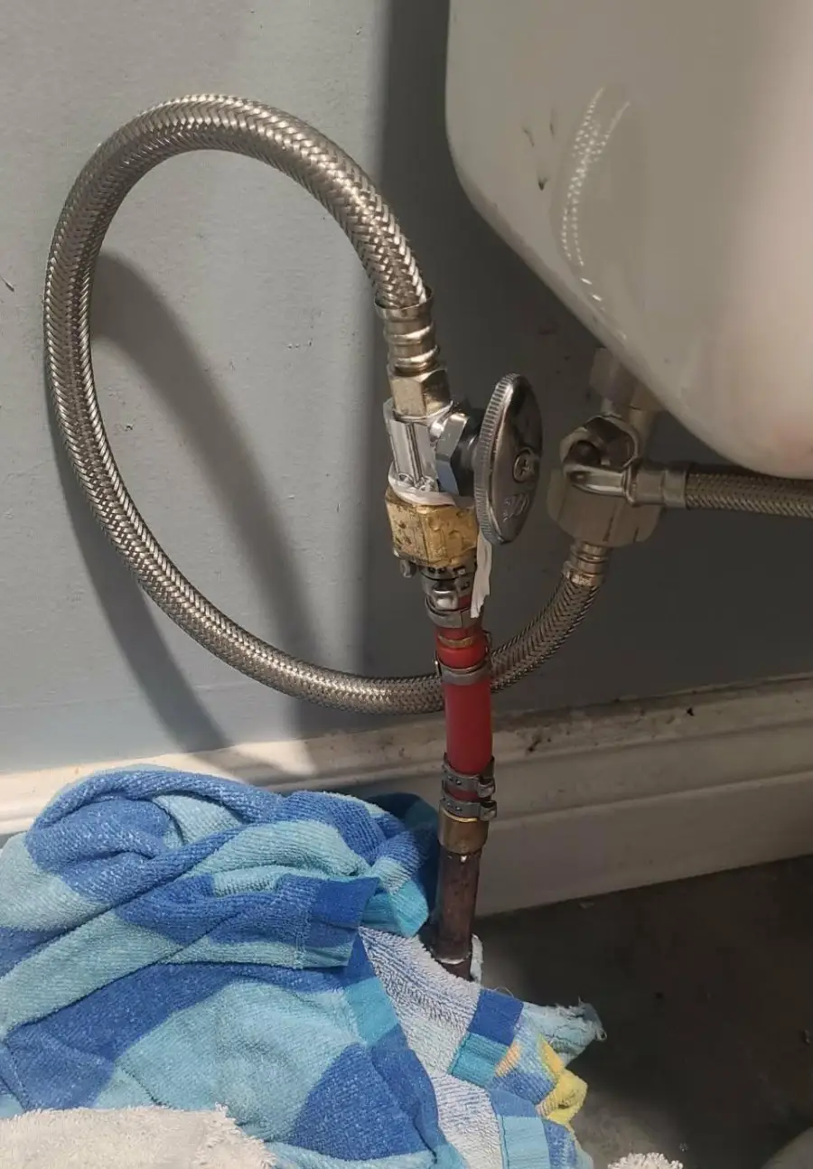 A makeshift toilet plumbing repair uses tape, a red pipe, and towels on the floor. Flexible hoses connect to the wall and toilet tank, with water stains visible on the baseboard.