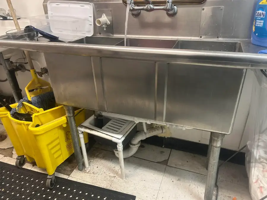 A stainless steel triple-compartment sink with cleaning supplies nearby, a mop bucket and mop to the left, and a blue cleaning bottle on the right. The floor is tiled and slightly worn.