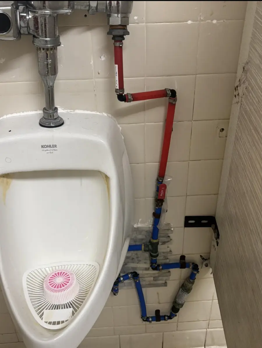 A white urinal is mounted on a tiled wall, with red and blue pipes running awkwardly along the wall and floor, leading to various connectors and valves. A pink urinal screen is visible inside the urinal.